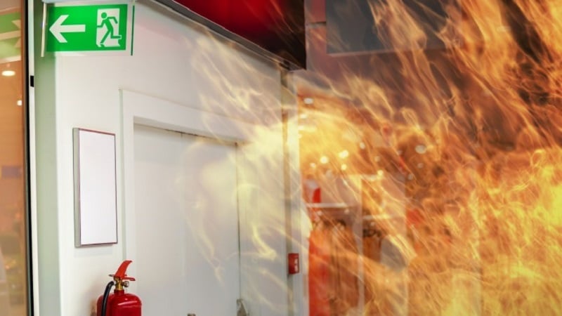 A fire in a room with an emergency exit sign and a fire extinguisher hanging on the wall.